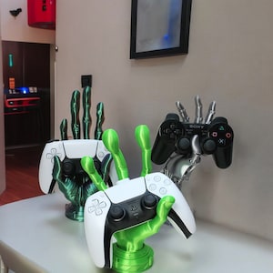 Könnte beinhalten: Zwei weiße PlayStation 5-Controller werden von 3D-gedruckten grünen Händen gehalten. Die Hände halten die Controller so, dass sie eine Person simulieren, die ein Videospiel spielt. Die Controller befinden sich auf einer weißen Oberfläche. Die Controller sind beide weiß mit schwarzen Knöpfen und einem schwarzen PlayStation-Logo. Die Hände sind grün und haben eine strukturierte Oberfläche. Die Hände halten die Controller so, dass sie eine Person simulieren, die ein Videospiel spielt.