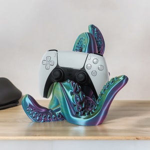 Könnte beinhalten: Ein weißer PlayStation 5-Controller sitzt in einem regenbogenfarbenen, lila und grünen, krakenförmigen Ständer. Der Ständer hat ein strukturiertes, organisches Design.