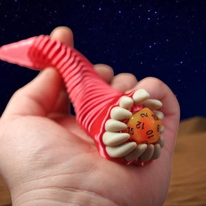 May include: A red, worm-like 3D-printed dice holder with a set of white teeth holding an orange D20 die. The background is a desert scene with a starry night sky.