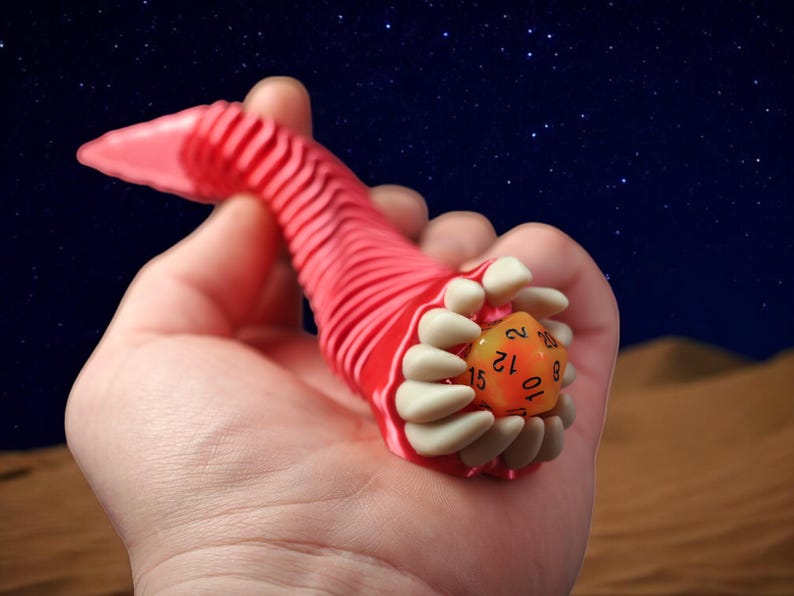May include: A red, worm-like 3D-printed dice holder with a set of white teeth holding an orange D20 die. The background is a desert scene with a starry night sky.