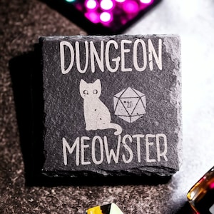 以下が含まれることがあります： 黒いスレートコースターに白い文字で「Dungeon Meowster」と書かれています。コースターには、白い漫画の猫と20面ダイスも描かれています。