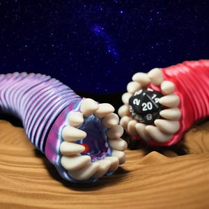 May include: Two stylized, worm-like creatures with open mouths and rows of teeth. One is pink and purple, the other is red, each holding a black, twenty-sided die. The background features a starry night sky and a sandy surface.
