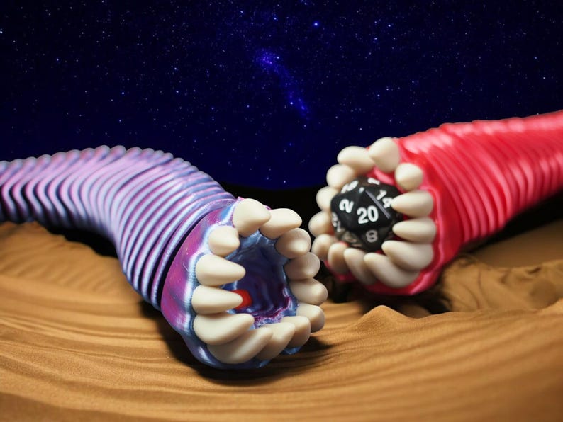 May include: Two stylized, worm-like creatures with open mouths and rows of teeth. One is pink and purple, the other is red, each holding a black, twenty-sided die. The background features a starry night sky and a sandy surface.
