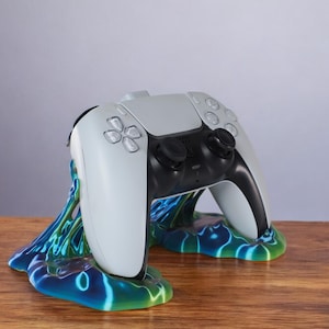 May include: A white video game controller with black buttons and a black handle is sitting on a blue, green and black abstract stand. The stand is on a wooden surface.