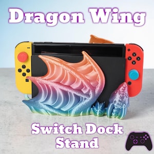 May include: A Switch dock stand with a dragon wing design. The stand is multi-coloured, with shades of orange, pink, blue, and purple. The text "Dragon Wing" and "Switch Dock Stand" are displayed on the image. A game controller icon is also present.