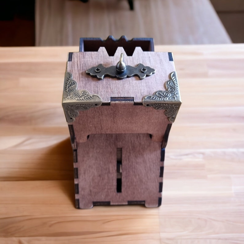 May include: A wooden box with a hinged lid and decorative metal corner accents. The box has a brass-colored handle and a decorative metal plate on the lid. The box is made of light brown wood and has a slot on the front.