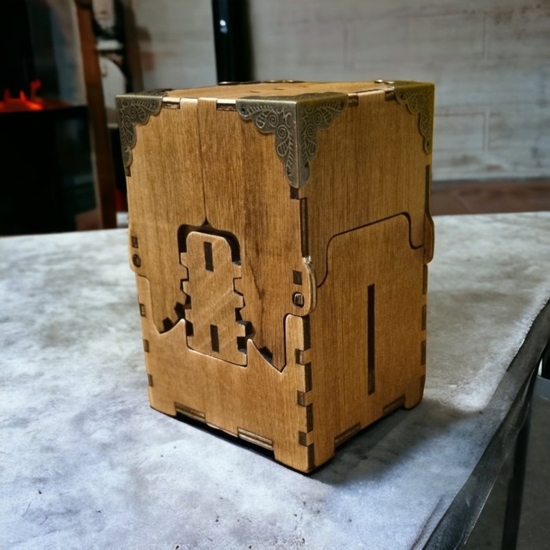 May include: A wooden box with a cube shape, featuring decorative metal corner accents. The box has a light brown color and a geometric design cut into the side. The box is sitting on a gray surface.