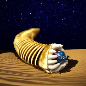 May include: A gold-colored, segmented, curved sculpture with white claw-like hands holding a blue and purple 20-sided die. The background features a starry night sky and a sandy surface.