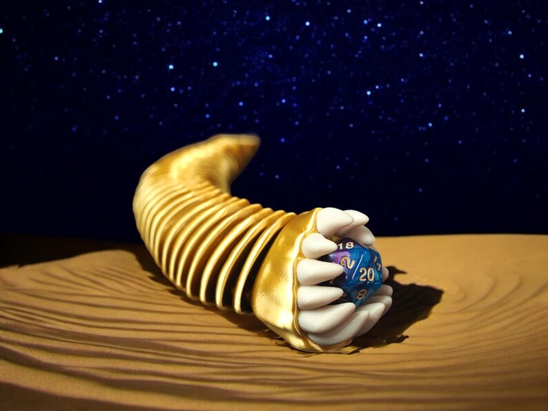 May include: A gold-colored, segmented, curved sculpture with white claw-like hands holding a blue and purple 20-sided die. The background features a starry night sky and a sandy surface.