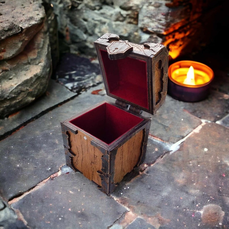 May include: A small, open wooden box with a hinged lid. The box is brown with dark brown trim and a red velvet interior. The lid is decorated with carved details. A lit candle is in the background.