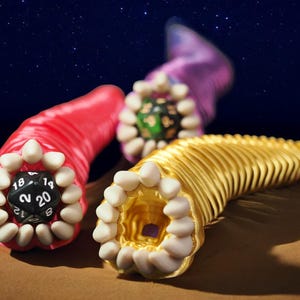 May include: Three colorful, worm-like sculptures with open mouths and teeth. One is red with a black die showing the number 20. Another is gold, and the third is purple. The background is a starry night sky.