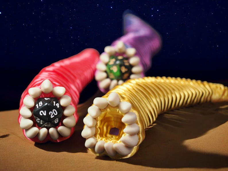 May include: Three colorful, worm-like sculptures with open mouths and teeth. One is red with a black die showing the number 20. Another is gold, and the third is purple. The background is a starry night sky.