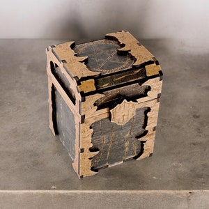 May include: A wooden box with a dark gray and tan finish. The box has intricate cut-out designs and a pentagram on the top. The box appears to be a puzzle box or a decorative storage container.