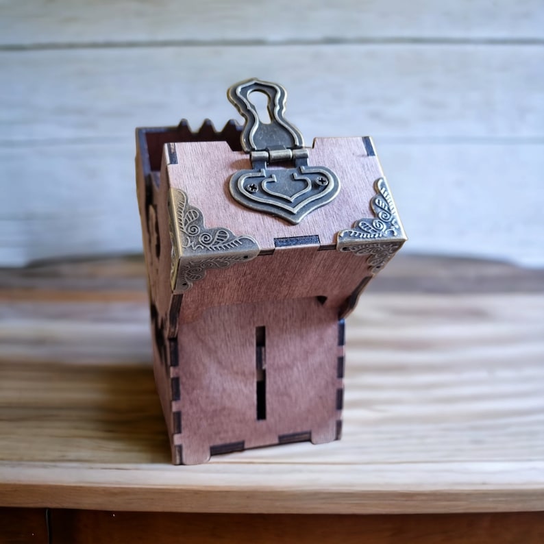 May include: A small, wooden box with a hinged lid and decorative metal hardware. The box is made of light brown wood and features ornate corner accents and a clasp. The box has a slot on the front and is sitting on a wooden surface.