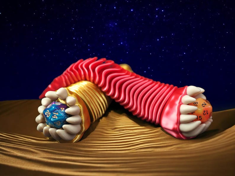 May include: A whimsical, fantasy-themed sculpture featuring a red, ridged form with gold accents, holding two multi-sided dice. One die is blue, the other orange, set against a starry night sky and a textured, golden surface.