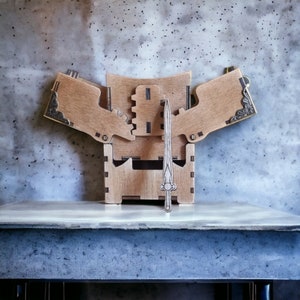 May include: A wooden box with a hinged lid and decorative metal corner accents. The box is open, revealing a wooden sword standing upright in the center. The box and sword are light brown, set against a gray background.