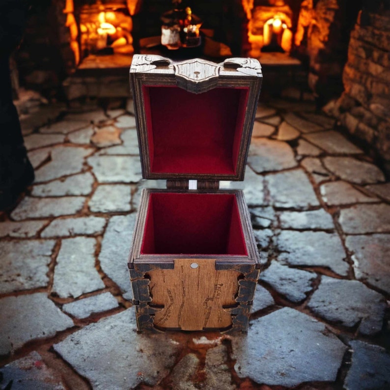 May include: Wooden treasure chest with an open lid, revealing a red velvet interior. The chest has intricate carved details and is set against a stone floor with a fireplace in the background. The chest is approximately 6 inches wide.