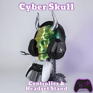 Puede incluir: Un soporte para mando y auriculares Cyber Skull. El soporte presenta una calavera verde con cuernos plateados, que sostiene unos auriculares negros. Un mando blanco y negro descansa en la base. Se muestra el texto "Cyber Skull" y "Controller & Headset Stand".