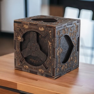 Puede incluir: Una caja de madera marrón oscuro con intrincados diseños cortados con láser. La caja tiene una forma geométrica tridimensional en el centro y una abertura en forma de corazón en la parte superior.