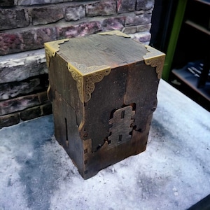May include: A dark brown wooden box with decorative brass corner accents. The box has a square shape and a detailed, carved design on the front. The wood grain is visible, giving it a rustic appearance. The box is sitting on a gray surface.