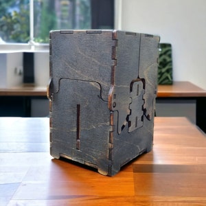 May include: A dark stained wooden box with a unique interlocking design. The box features a sliding lid and decorative cutouts on the side. The wood grain is visible, adding texture to the piece. The box is sitting on a wooden table.