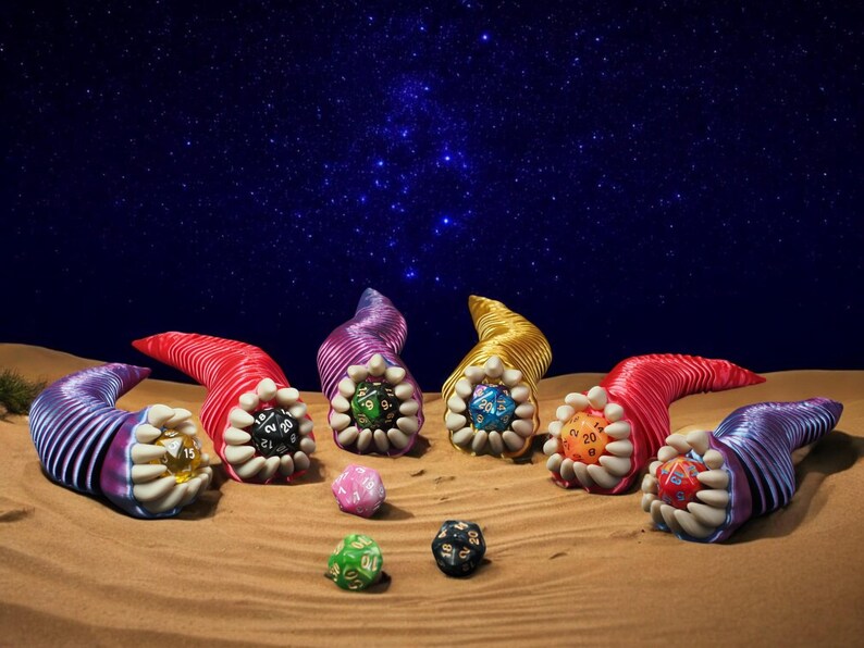 May include: A fantasy-themed image featuring colorful, horn-shaped objects with dice inside, set against a starry night sky. The horns are red, purple, and yellow, with white teeth-like structures. Several dice in various colors are scattered on the sand.