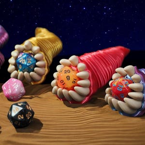 May include: An artistic composition featuring colorful, fantasy-themed dice. The dice are held in stylized, creature-like forms with open mouths and teeth. The background is a starry night sky, with the dice resting on a sandy surface.