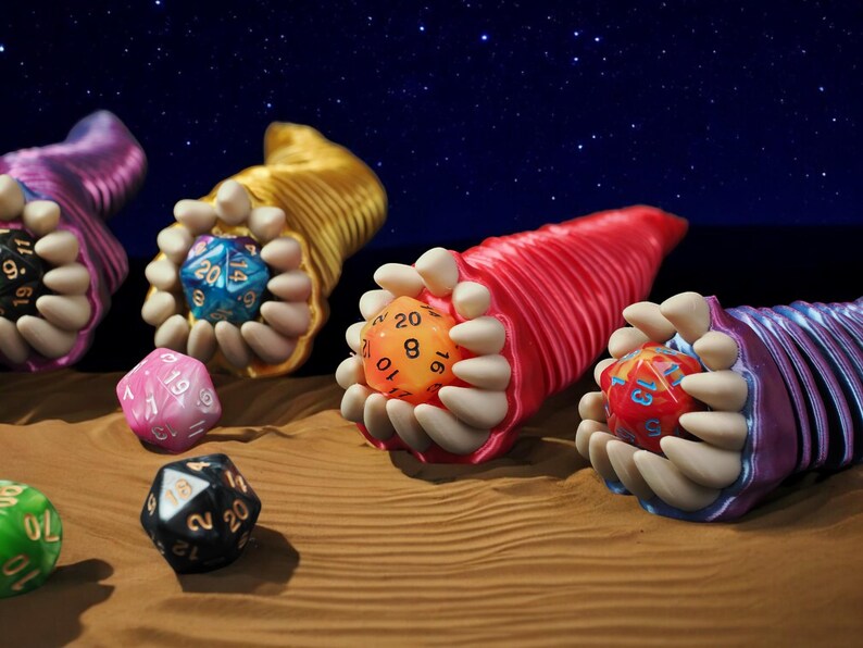 May include: An artistic composition featuring colorful, fantasy-themed dice. The dice are held in stylized, creature-like forms with open mouths and teeth. The background is a starry night sky, with the dice resting on a sandy surface.