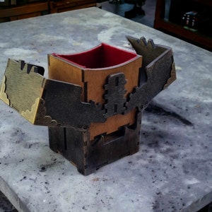 May include: A wooden box with a hinged lid, featuring intricate laser-cut designs and brass corner accents. The interior is lined with red fabric. The box is open, revealing the interior. The box is sitting on a gray surface.