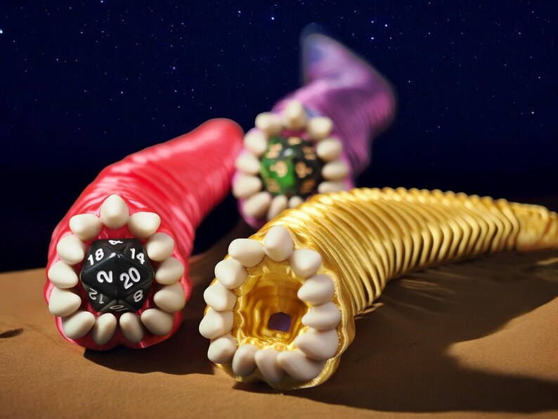 May include: Three colorful, stylized worm-like objects with open mouths and teeth. One is red with a black die showing the number 20. Another is gold, and the third is purple. The background is a starry night sky.