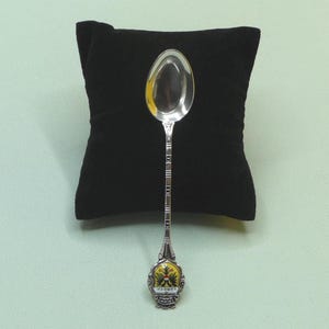 Puede incluir: Una cuchara plateada con un mango detallado y un cuenco decorativo. El mango presenta una serie de detalles rectangulares y cuadrados. El cuenco de la cuchara refleja el entorno. La base de la cuchara tiene un diseño amarillo y verde con la palabra "LUBECK".