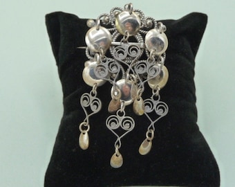 May include: A silver filigree brooch with a central round pendant and multiple dangling pendants. The brooch is displayed on a black velvet background.