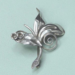 Peut inclure: Broche argentée au design floral. La broche présente des feuilles stylisées, deux petites fleurs et un élément enroulé. L'épingle est posée sur un fond bleu-vert clair.