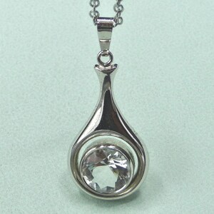 May include: A silver-toned pendant necklace featuring a teardrop-shaped design. The pendant has a clear, faceted gemstone set within a circular frame. The necklace chain is also silver-toned and delicate.