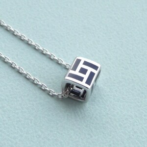 Peut inclure: Un collier en chaîne argent avec un pendentif en forme de cube noir et argent. Le cube a un motif géométrique.