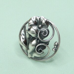 May include: A silver-toned brooch with a floral design. The brooch features a central flower with detailed petals, surrounded by leaves and a decorative circular frame. The design includes swirling accents and small, round embellishments.