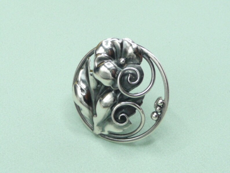 May include: A silver-toned brooch with a floral design. The brooch features a central flower with detailed petals, surrounded by leaves and a decorative circular frame. The design includes swirling accents and small, round embellishments.