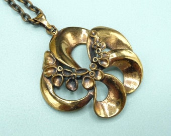 May include: A gold-toned pendant necklace featuring a stylised floral design. The pendant is made up of a series of overlapping, curved shapes with a textured surface.