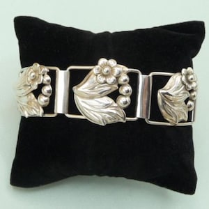 Peut inclure: Un bracelet en argent avec un motif floral. Le bracelet est composé de plusieurs sections reliées, chacune présentant une fleur stylisée avec des feuilles.