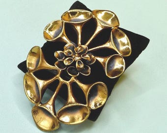 Peut inclure: Broche en métal doré avec un motif floral abstrait. La broche présente un groupe central de petites formes arrondies entourées de plus grands éléments en forme de pétales. La broche est présentée sur une surface en velours noir.