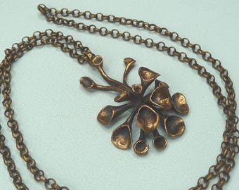 May include: A bronze-toned necklace featuring a pendant with an organic, floral-like design. The pendant has multiple rounded, petal-shaped elements. The chain is a matching bronze color, with a classic link style. The necklace rests on a light blue surface.