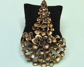 May include: A large, bronze-colored pendant with a unique, organic design. The pendant features numerous rounded, cup-shaped elements connected by thin stems, creating a clustered, tree-like form. The pendant is displayed on a black velvet surface.