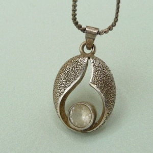May include: Silver-toned pendant necklace featuring an oval-shaped pendant with a textured surface and a teardrop-like cutout. A clear, round stone is set at the bottom of the pendant. The necklace has a delicate chain.