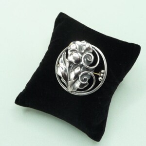 May include: A silver-toned floral brooch with a detailed design of leaves and a flower, set within a circular frame. The brooch is displayed on a black velvet pillow against a light green background.