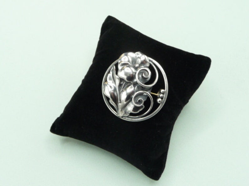 May include: A silver-toned floral brooch with a detailed design of leaves and a flower, set within a circular frame. The brooch is displayed on a black velvet pillow against a light green background.