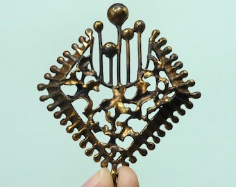 May include: A gold-toned metal brooch with a geometric design. The brooch features a central square shape with a raised, intricate pattern of branches and leaves. The square is surrounded by a border of small, rounded points.