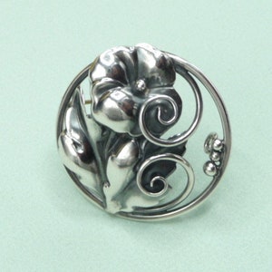 May include: A silver-toned floral brooch with a detailed flower and leaves design. The brooch features a circular frame with decorative swirls and small bead accents, creating an elegant, nature-inspired accessory.