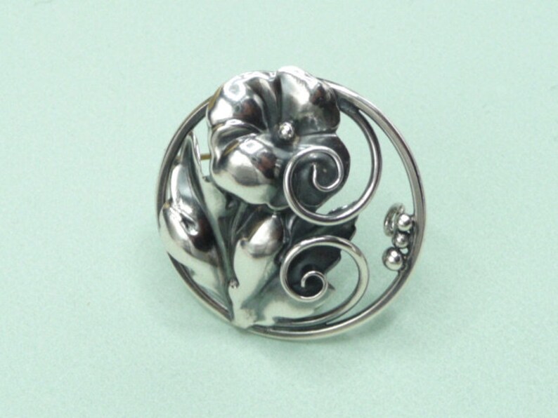 May include: A silver-toned floral brooch with a detailed flower and leaves design. The brooch features a circular frame with decorative swirls and small bead accents, creating an elegant, nature-inspired accessory.