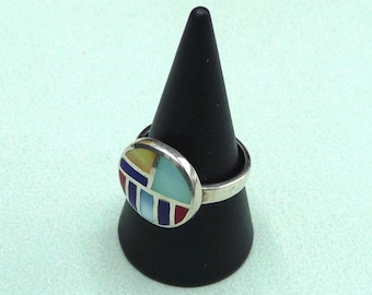 Puede incluir: Un anillo de plata con una incrustación colorida de piedras azules, rojas, amarillas y verdes. El anillo está en un expositor de anillos negro.