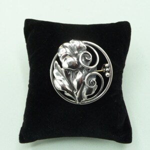 May include: A silver-toned floral brooch with a flower and leaf design, set within a circular frame. The brooch is displayed on a black velvet pillow against a light green background. The brooch has a pin closure on the back.
